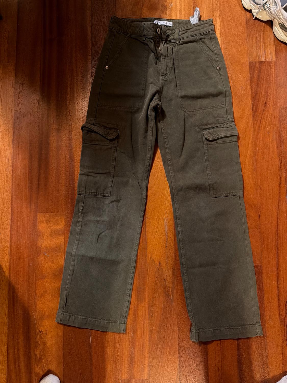Olive green cargo trousers on a wooden floor. Side pockets and visible tag on the waistband. Part of some white trainers on the right.