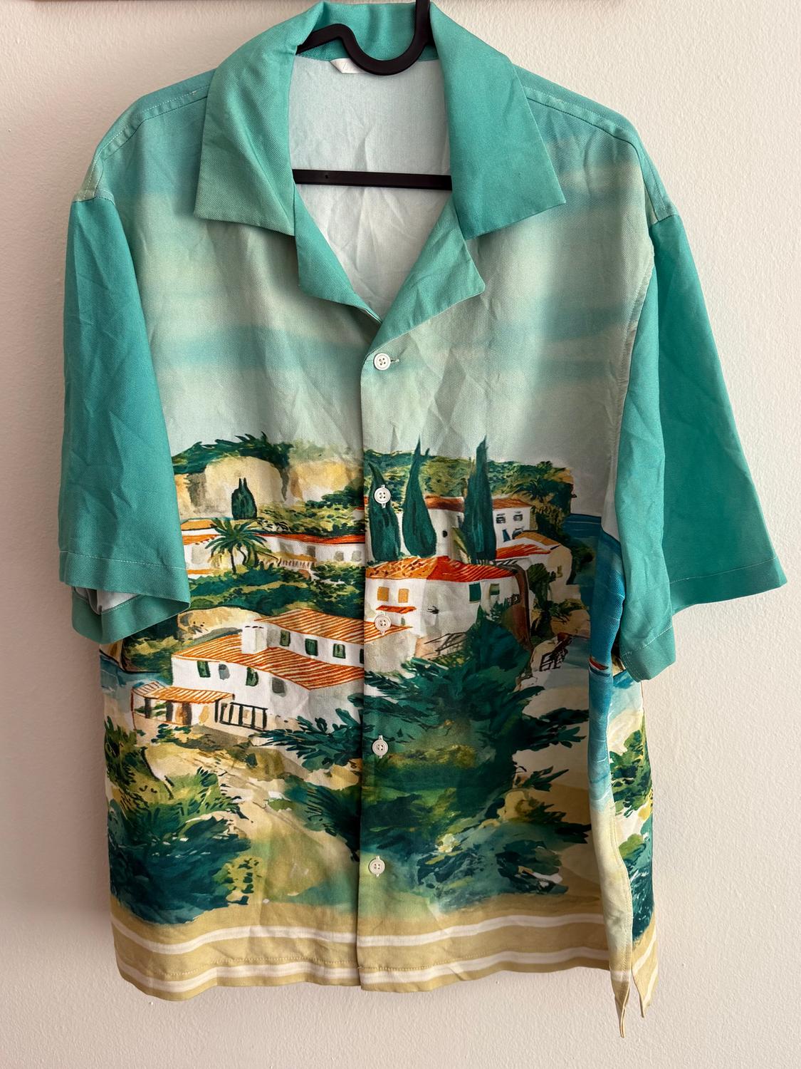 Short-sleeved shirt with Mediterranean landscape print, in green and blue tones, hanging on a hanger against a white wall.