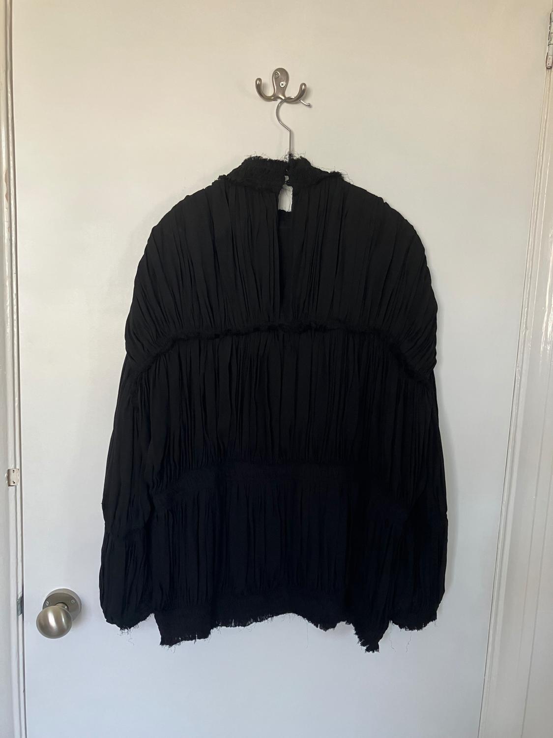 Black crinkled fabric blouse hanging on a white door. The blouse has a loose design and long sleeves.