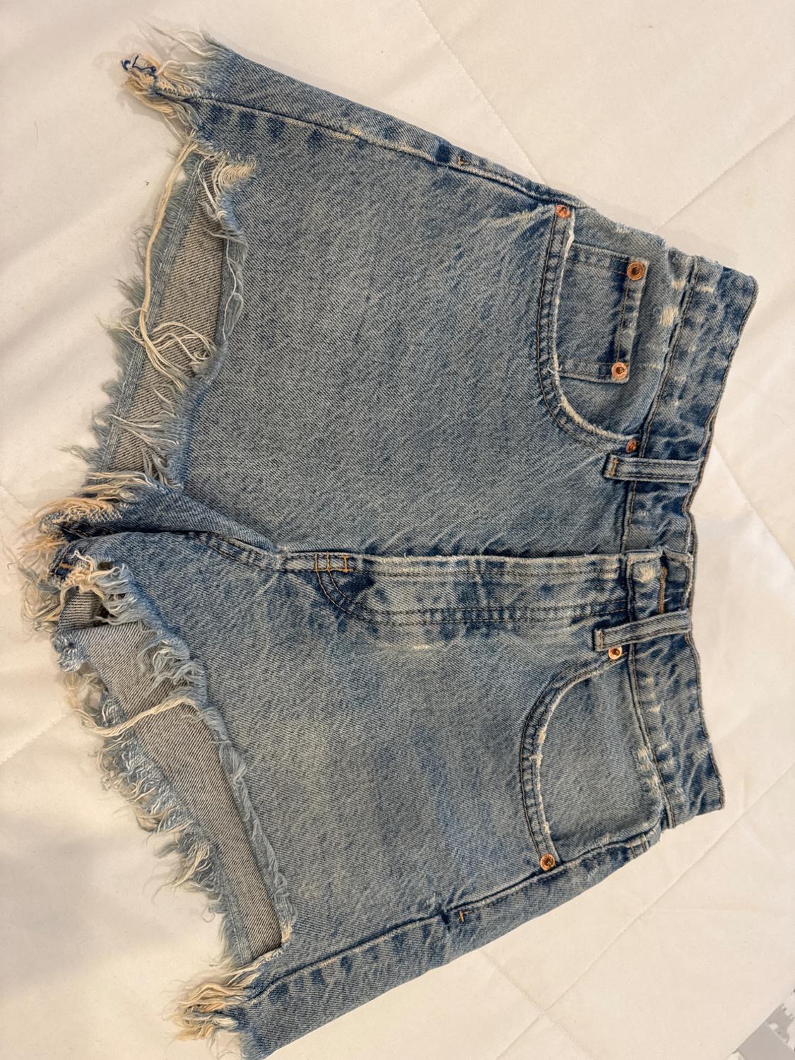 Light blue denim shorts with frayed edges on a white surface. Classic design with pockets and copper buttons.