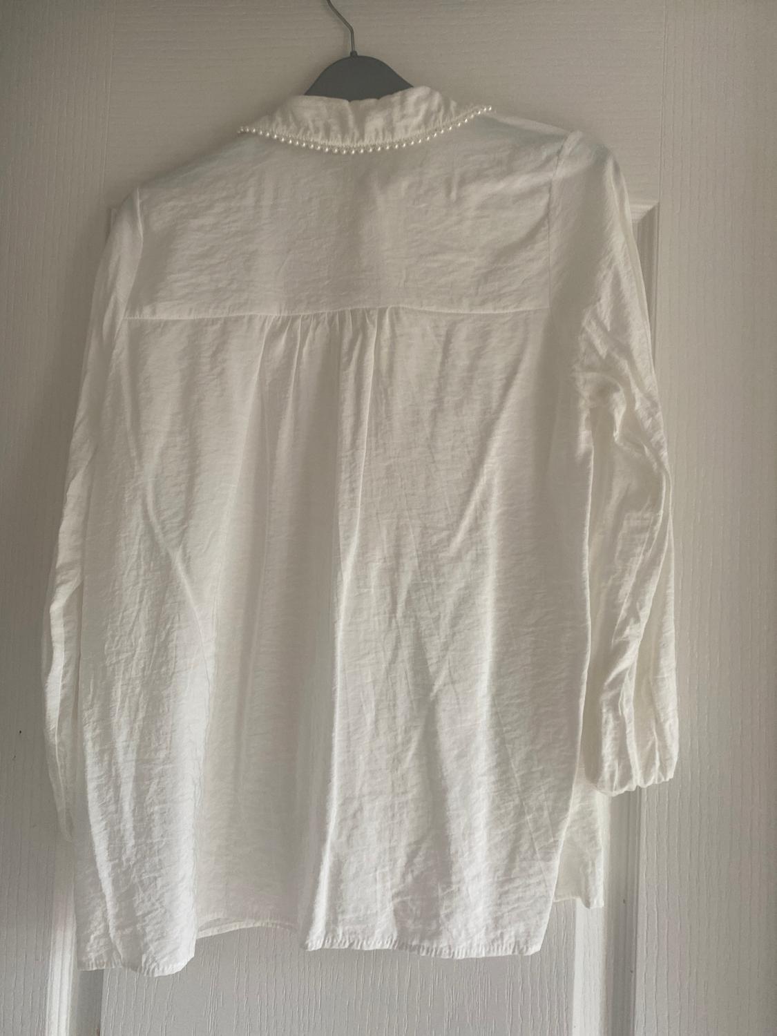 White long-sleeved shirt with pearl detailing on the collar, hanging on a hanger on a white door.