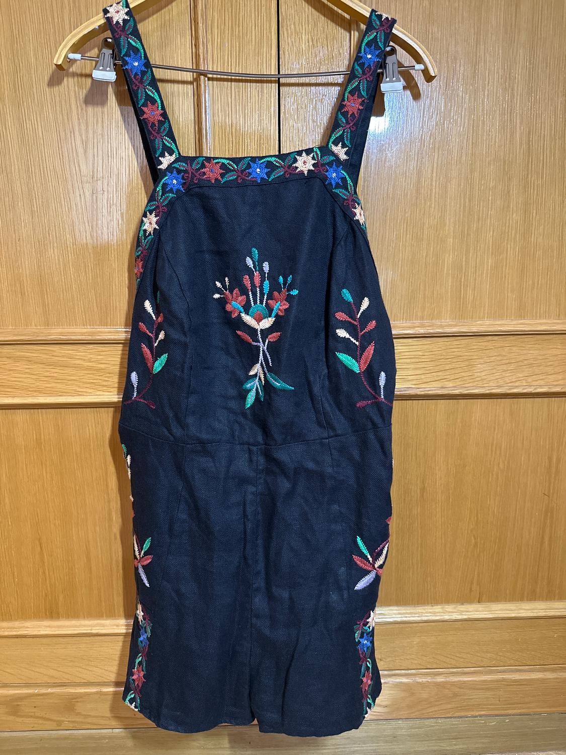 Black dress with straps, multi-coloured floral embroidery on the chest and sides. Hanging on a wooden hanger against a light wooden background.