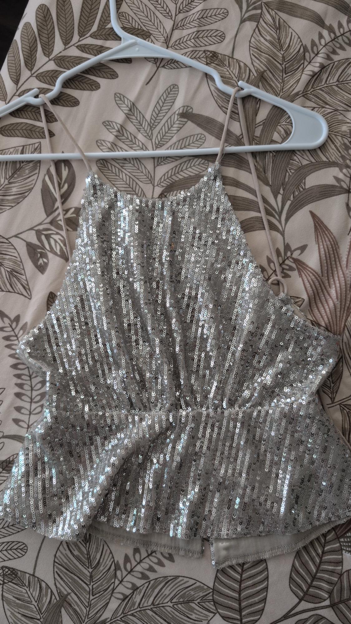 Silver sequin spaghetti strap top hanging on a white hanger against a leaf-print fabric.