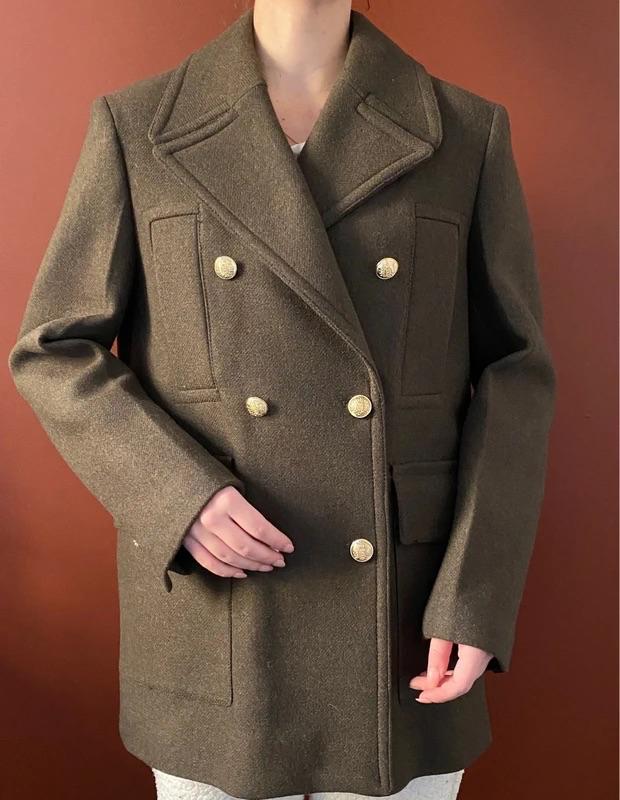 Model wearing an olive green double-breasted coat with gold buttons, against a brown background.