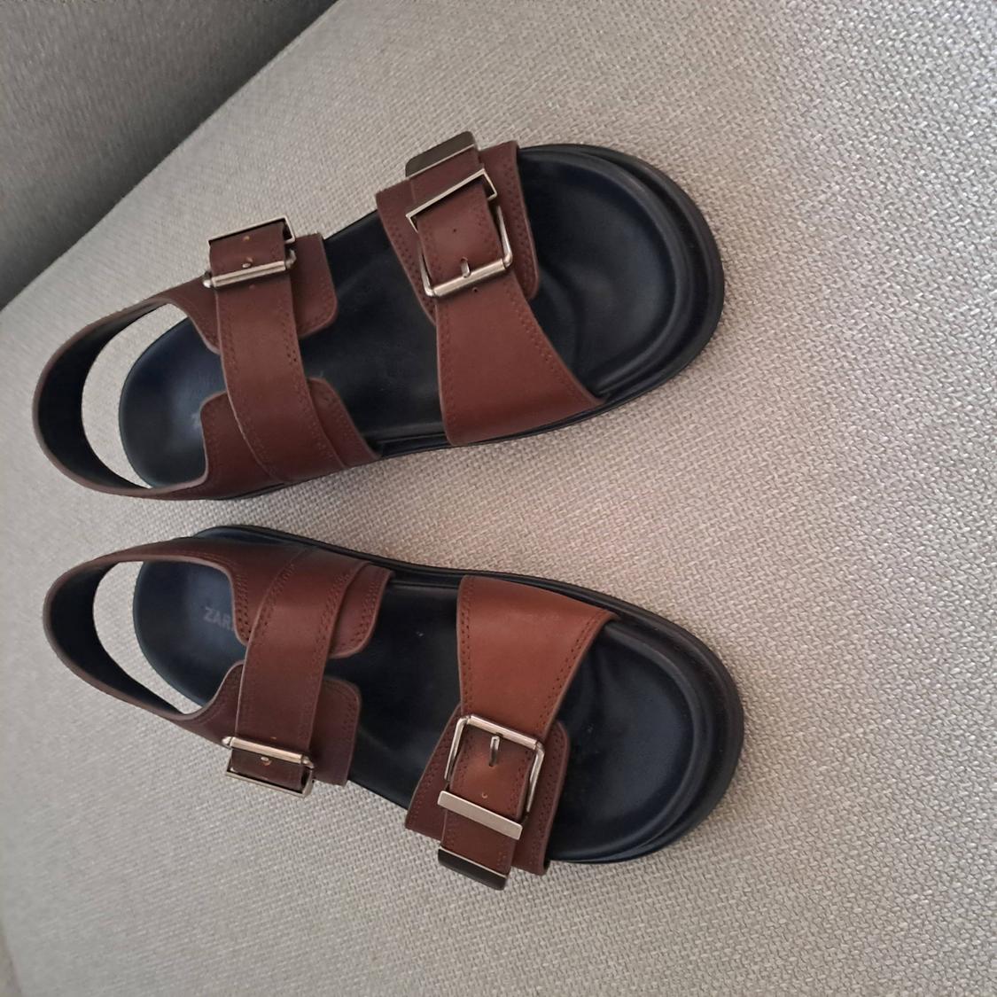 Brown sandals with two wide straps, silver buckles, and a black sole, resting on a textured grey fabric surface.