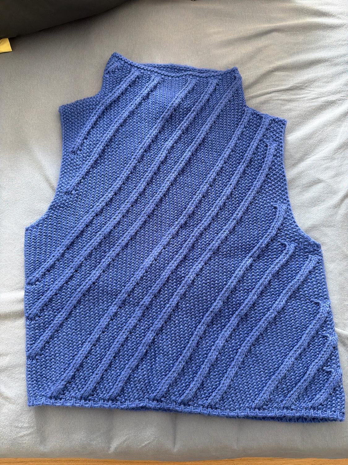 Blue sleeveless knit top with a high neck and raised diagonal lines, laid out on a crumpled light grey fabric surface.