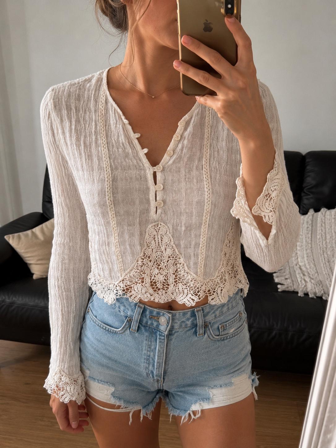 Woman taking a selfie. She is wearing a long-sleeved white blouse with lace and buttons, and distressed denim shorts. Black sofa in the background.