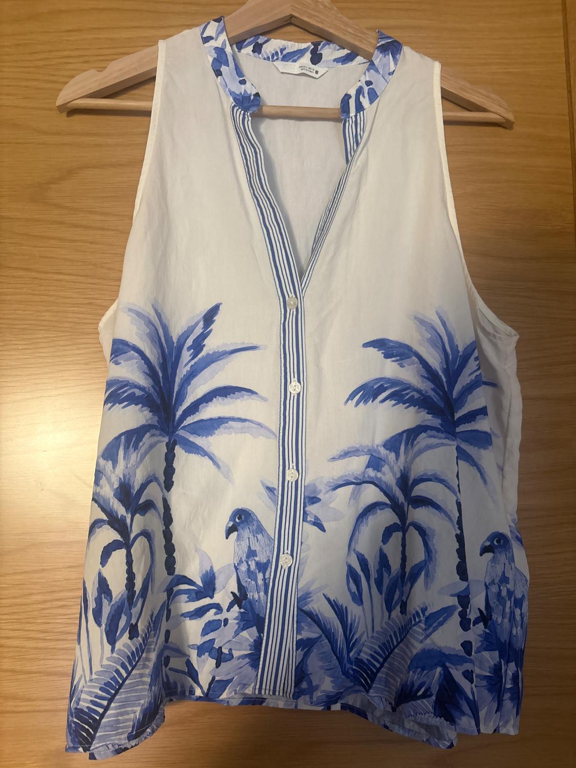 Sleeveless white blouse on a wooden hanger. Blue palm tree and parrot print, V-neck with striped trim and front buttons.