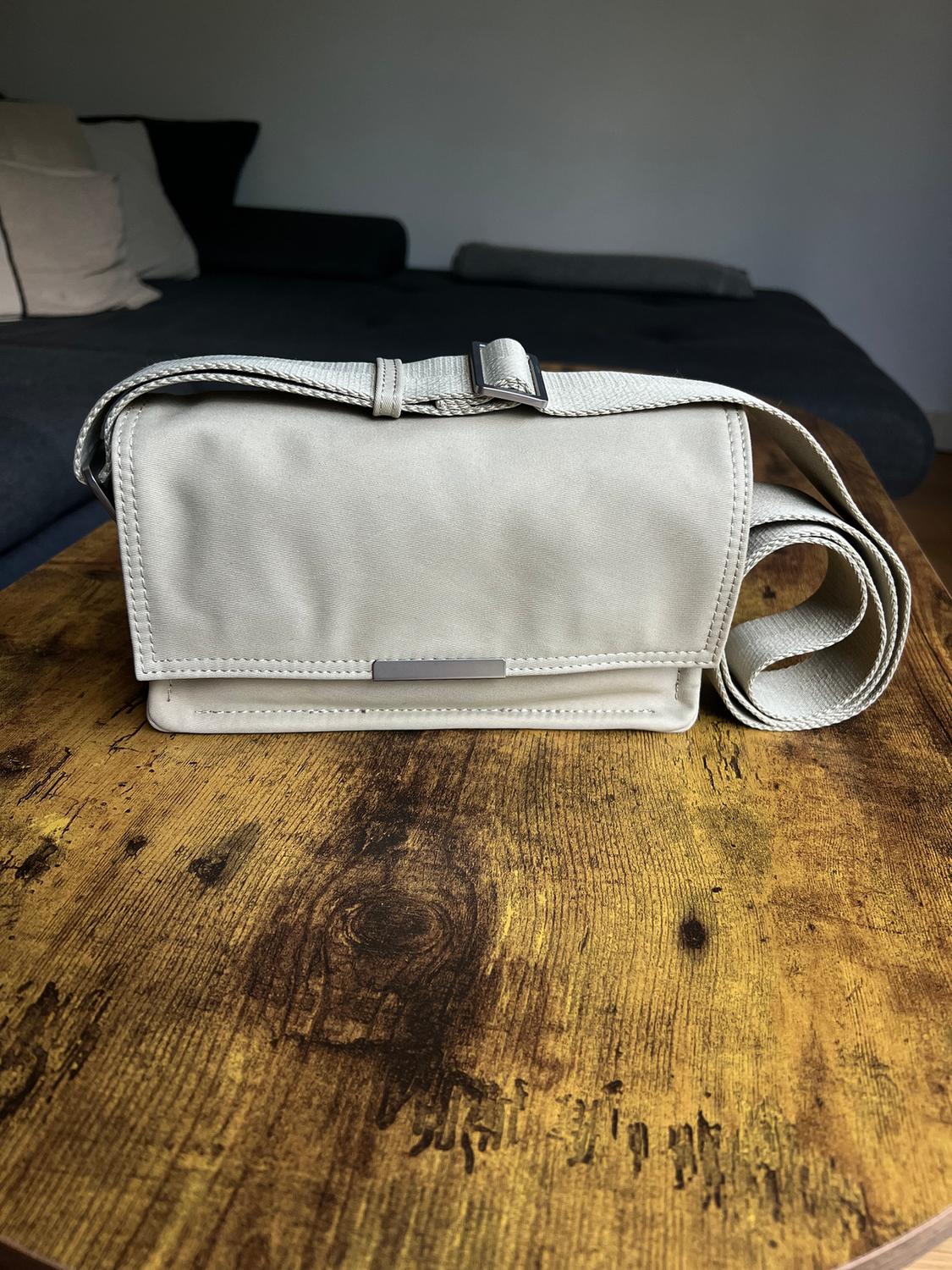 Beige handbag on wooden table, with adjustable strap and metal buckle. Background with dark sofa and light cushions.