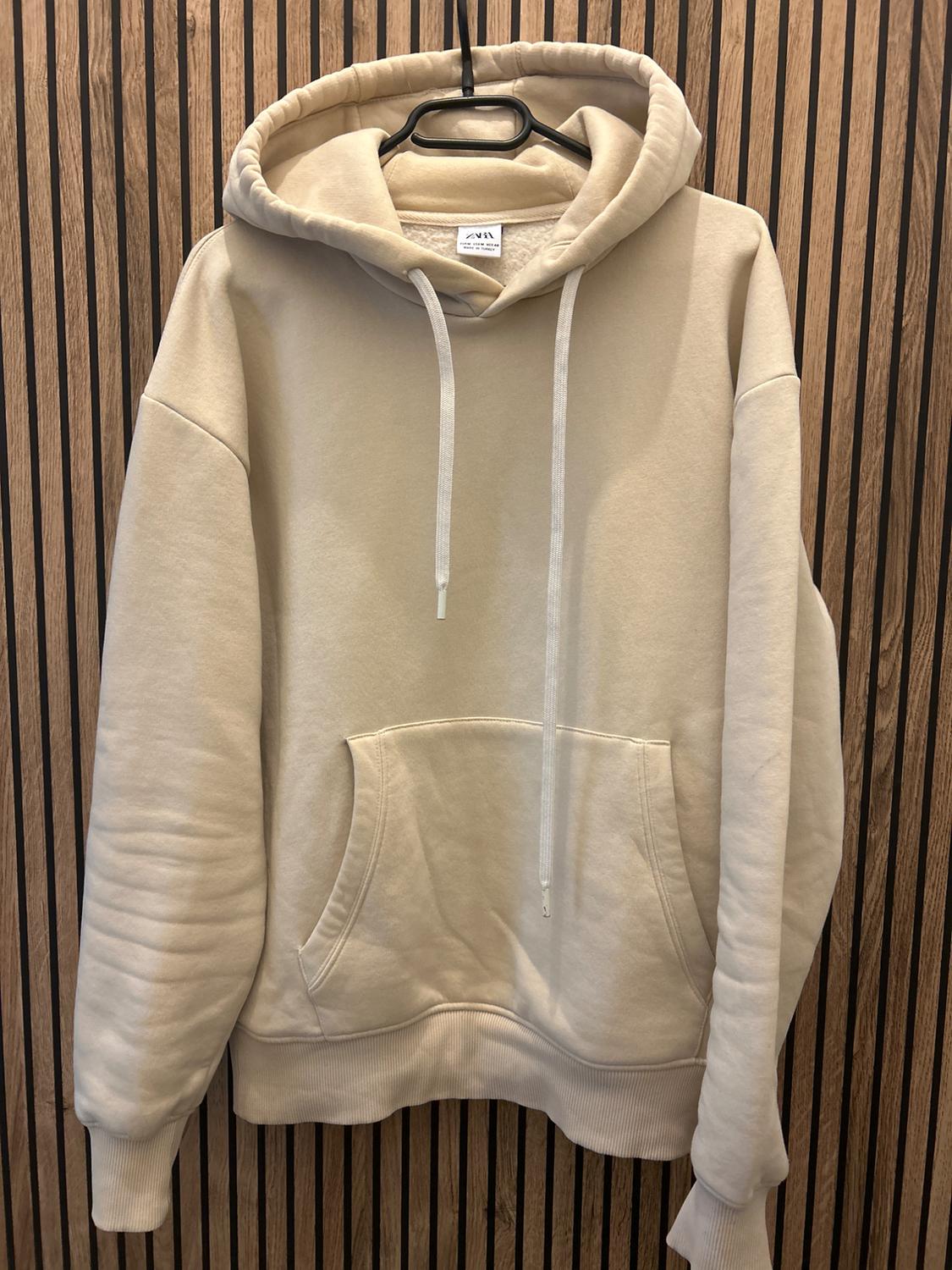 Beige hoodie with white drawstrings hanging on a black hanger, vertical wooden panel background.