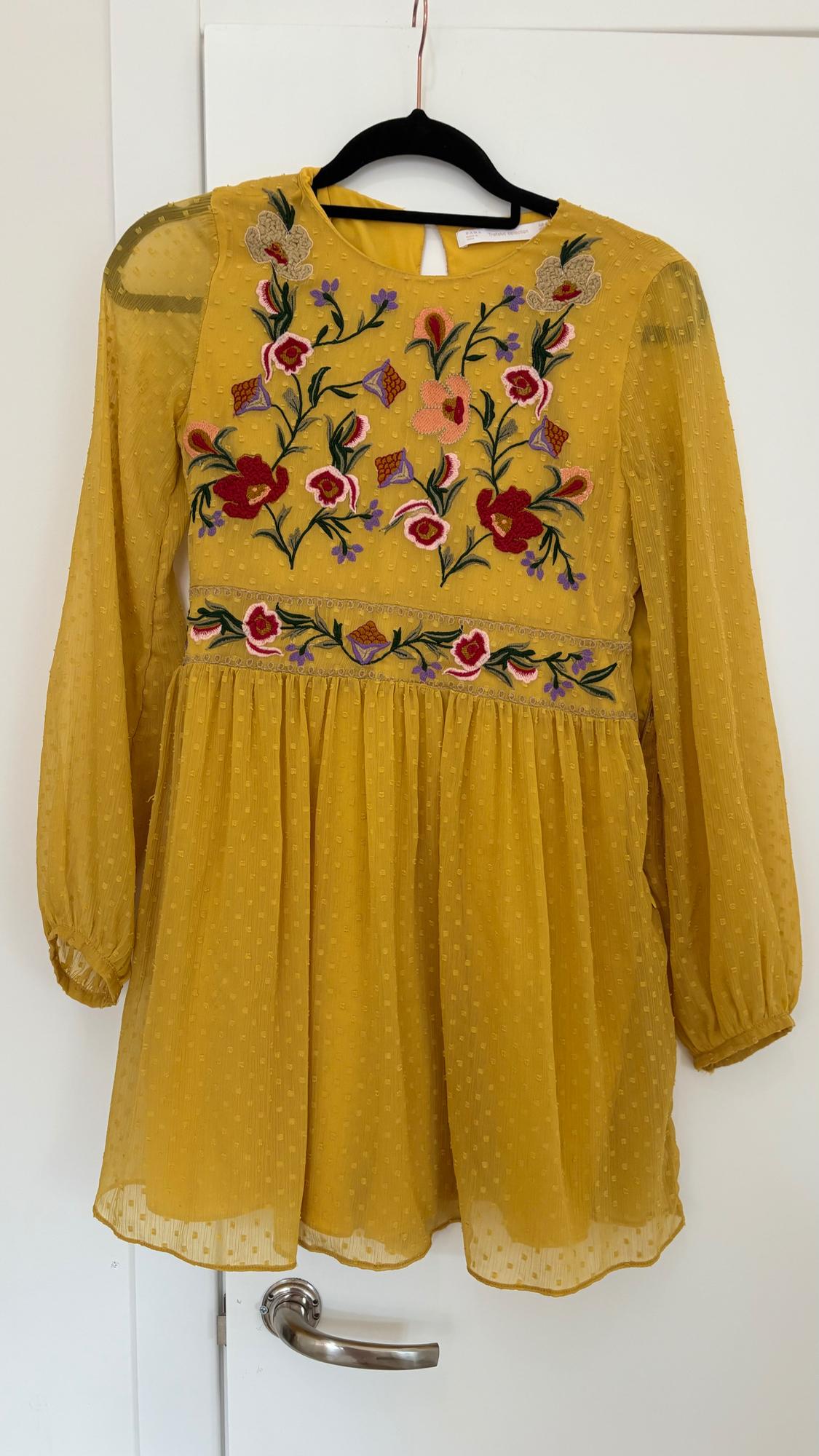 Yellow dress with long sleeves and floral embroidery in red, pink, and green. Hanging on a black hanger over a white door.