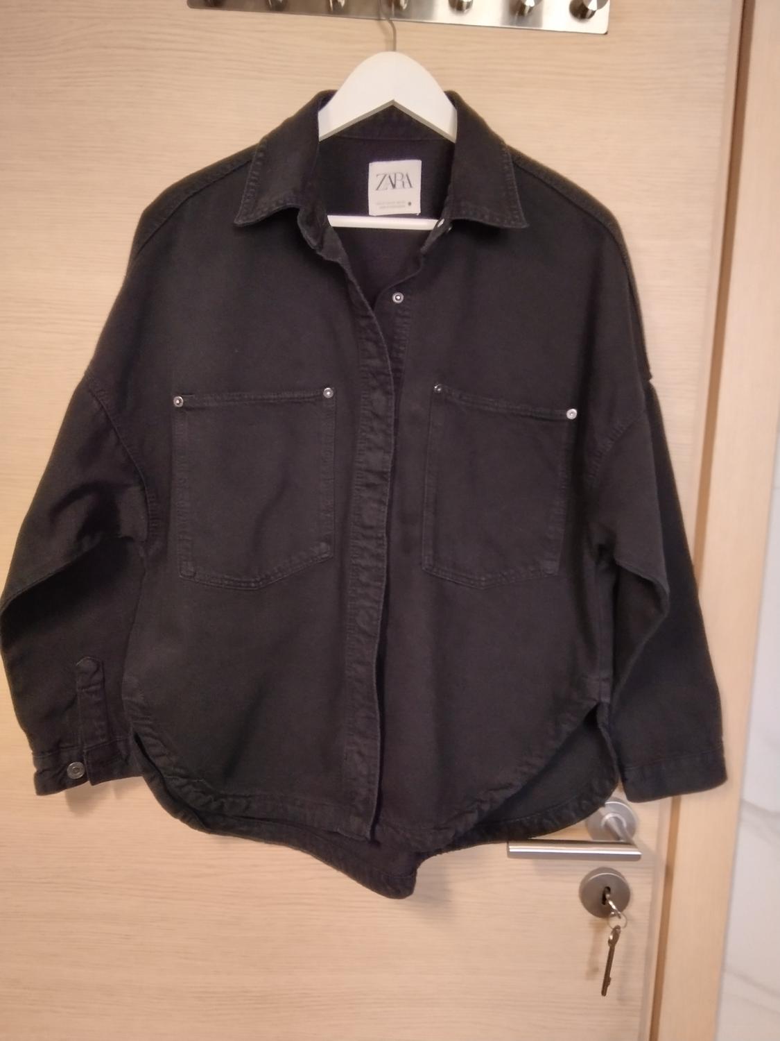 Long-sleeved black shirt with front pockets hanging on a white hanger over a wooden door. Label with the text ZARA.