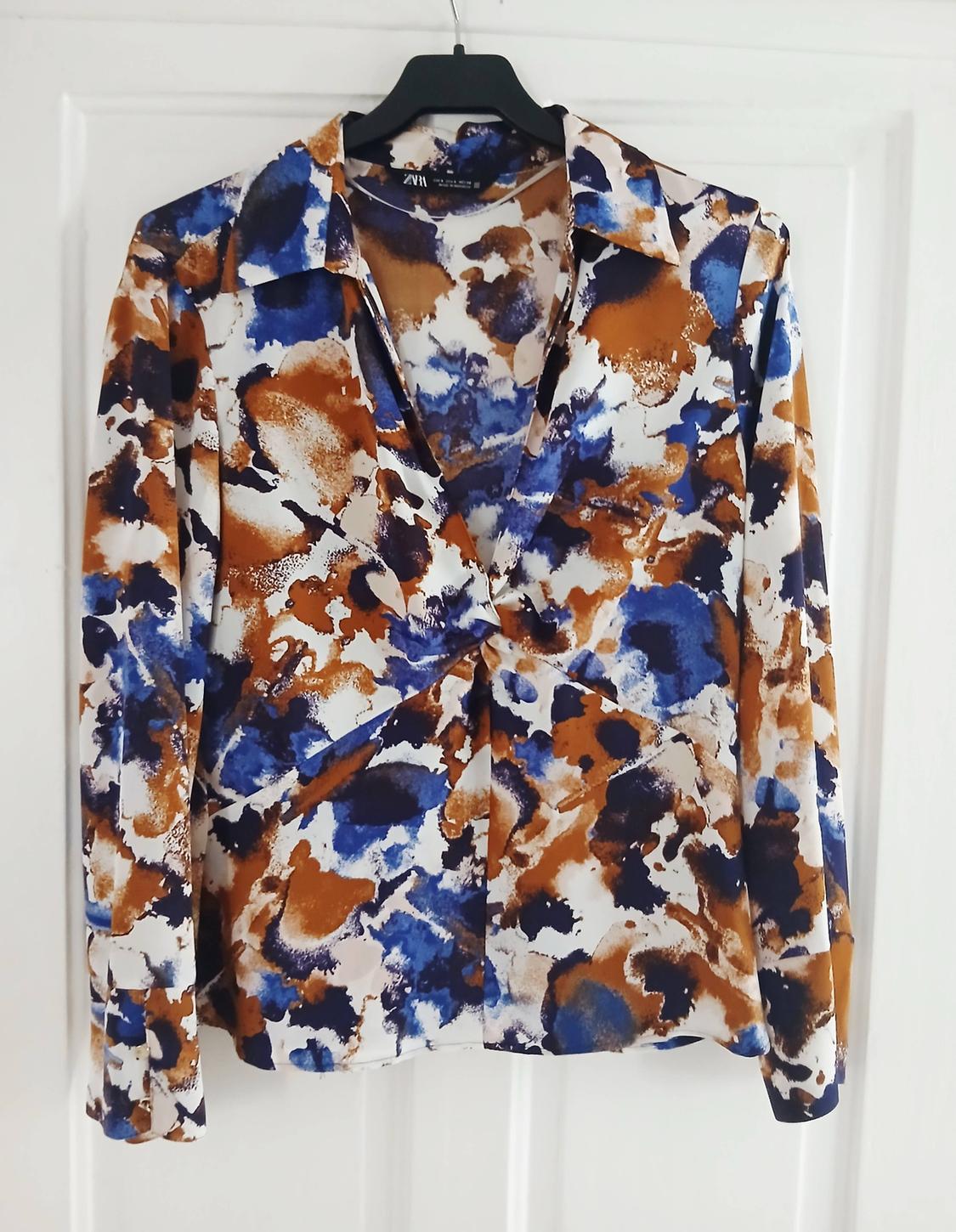 Printed blouse with abstract brown, blue, and white splodges, hanging on a black hanger in front of a white door.