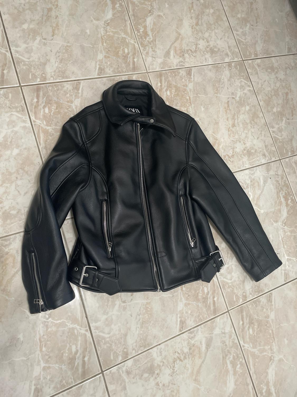 Black jacket with silver zips and buckles at the hem, on a tiled floor. ZARA label inside.