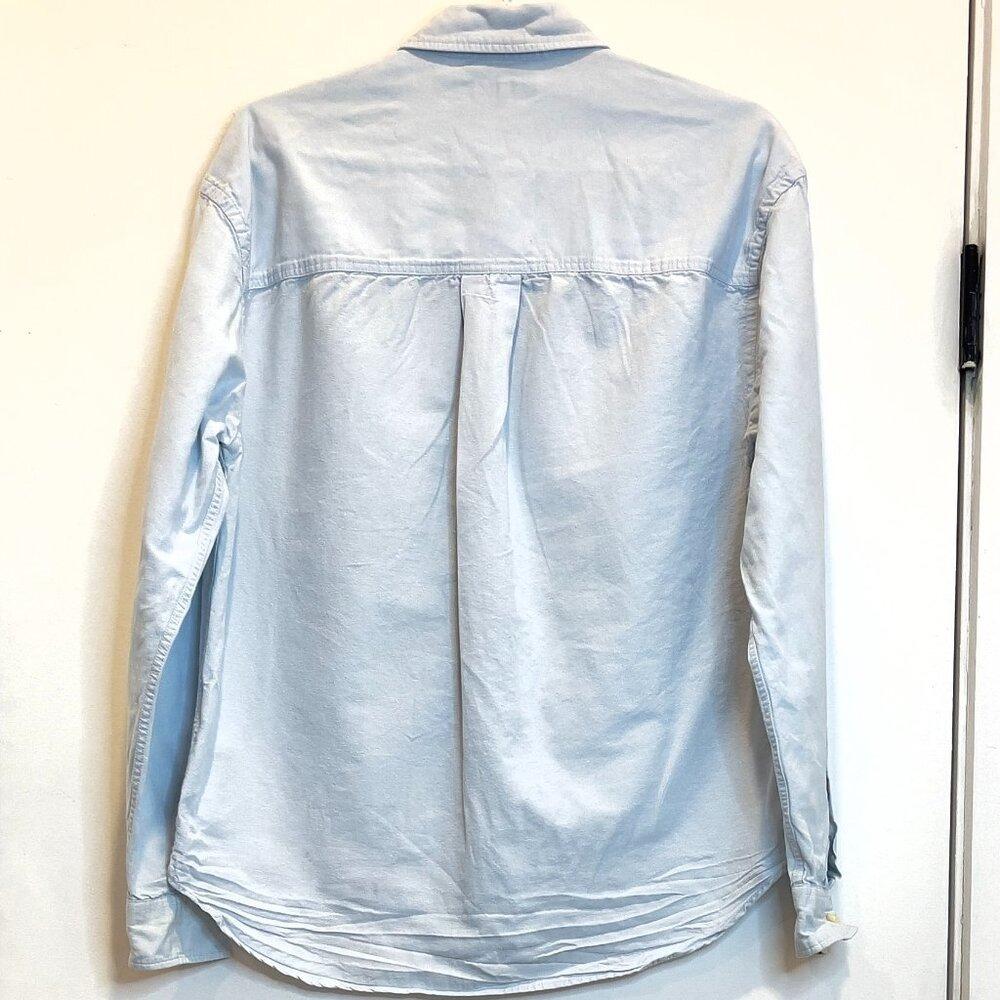 Back view of a light blue long-sleeve shirt with a collar and a central pleat, hanging on a plain white background.