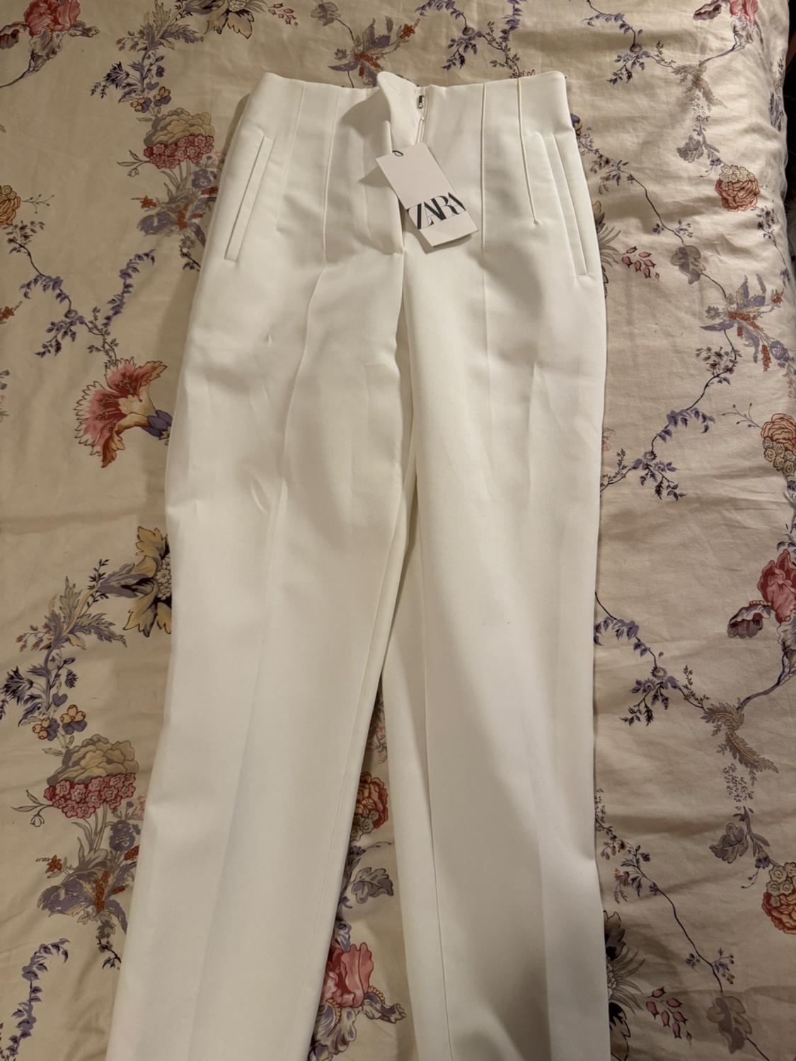 White pants with a "ZARA" tag on a floral bedspread in beige, red, and purple tones.
