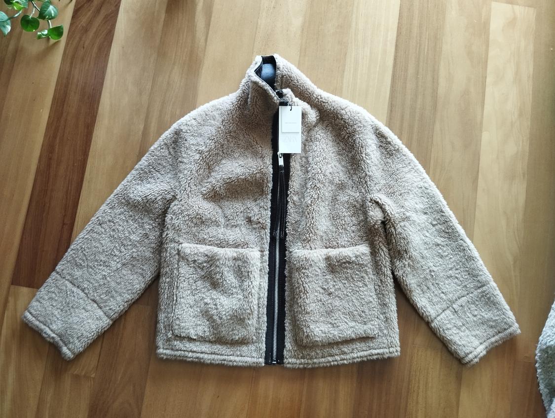 Beige fleece jacket with black zip and front pockets, on a light wooden floor. White tag hanging from the collar.