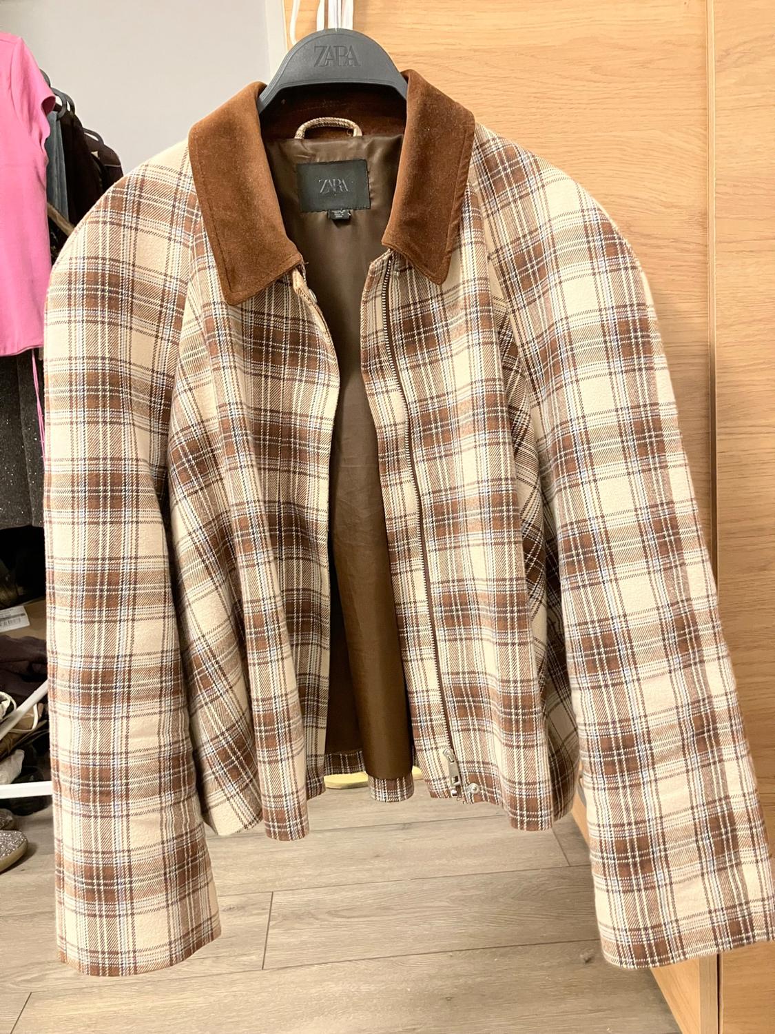 Beige and brown checked jacket with a brown collar hanging on a hanger with the text ZARA in a room with a wooden floor.