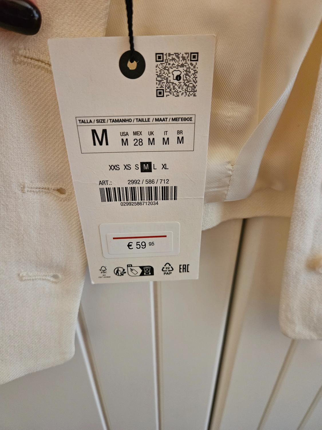 White clothing label with a price of €59.95. Available in size M in various regions. Barcode and QR visible. White wooden background.