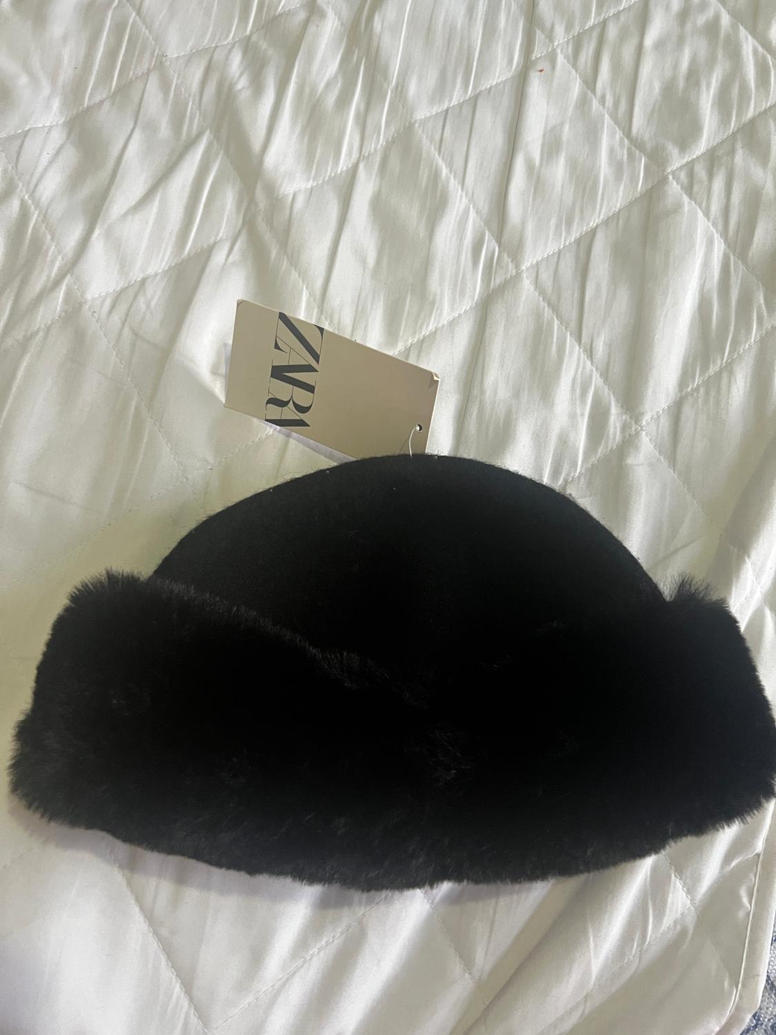 Black faux leather hat on a quilted white bedspread. Visible label with text "ZARA".