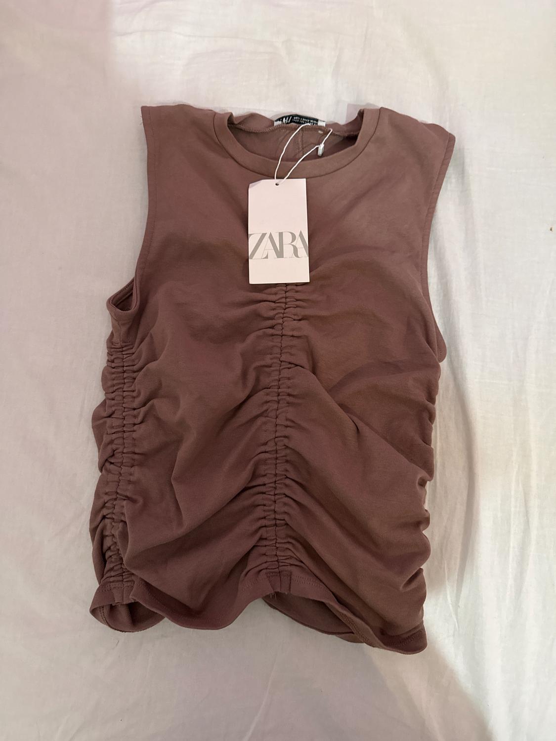 Brown sleeveless top with ruching at the center, hang tag with the text "ZARA", on a white background.