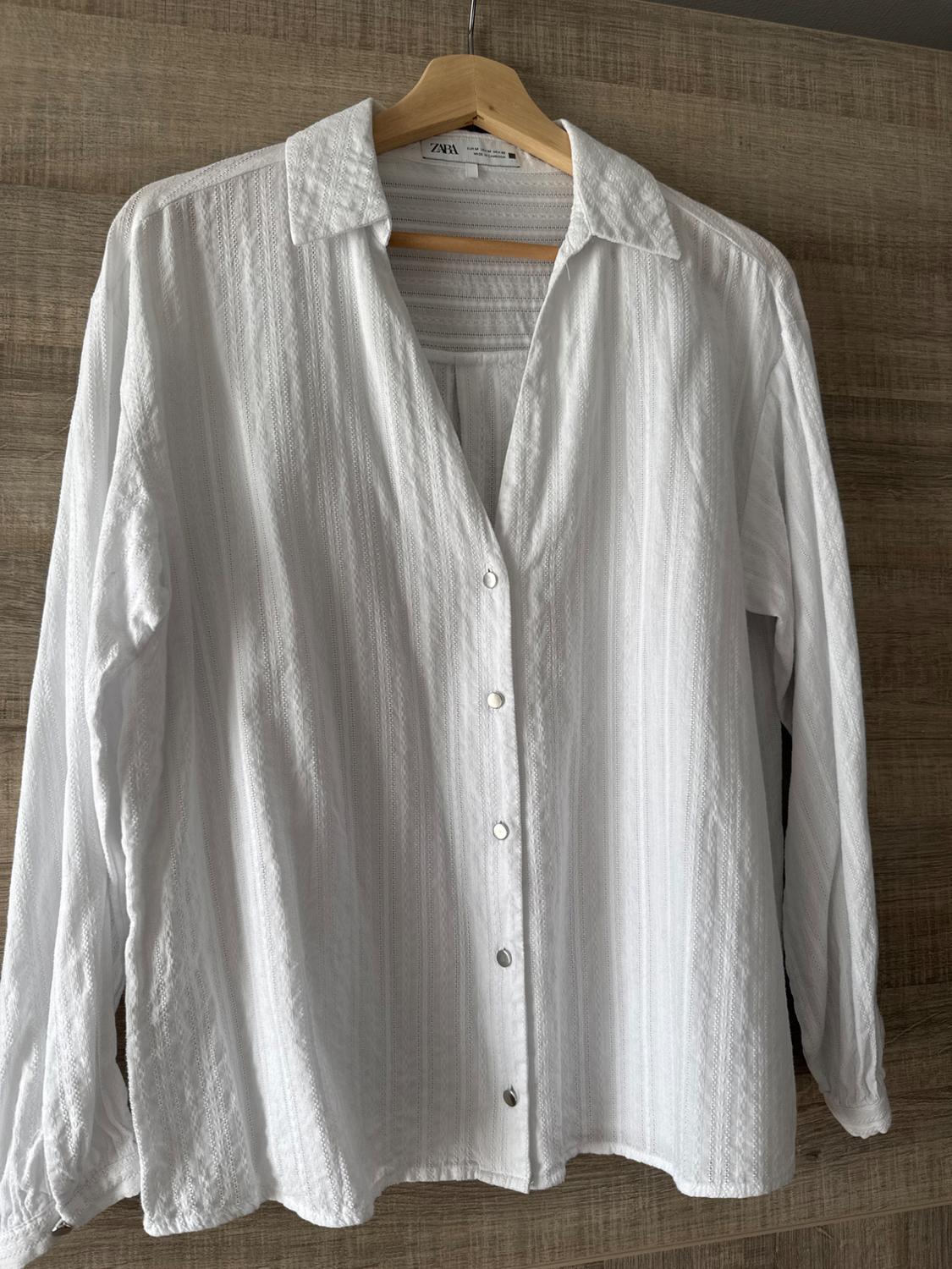 White long-sleeved button-up shirt, hanging on a wooden hanger against a light wood background. Visible tag.