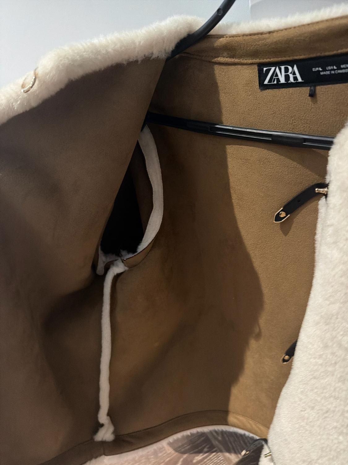 Detail of brown jacket with white lining on a black hanger. Interior label with the text ZARA and black straps with gold buckles.