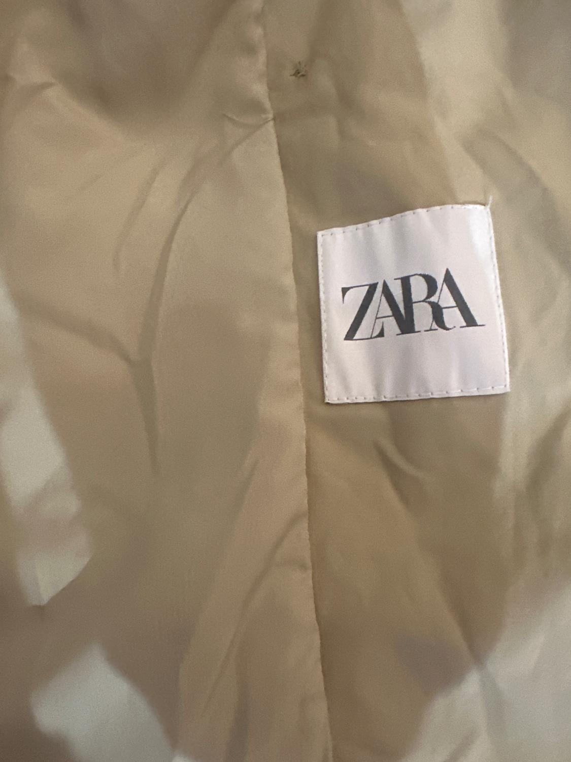 Close-up of a beige fabric with a central seam and a sewn-on square white label showing the text ZARA.