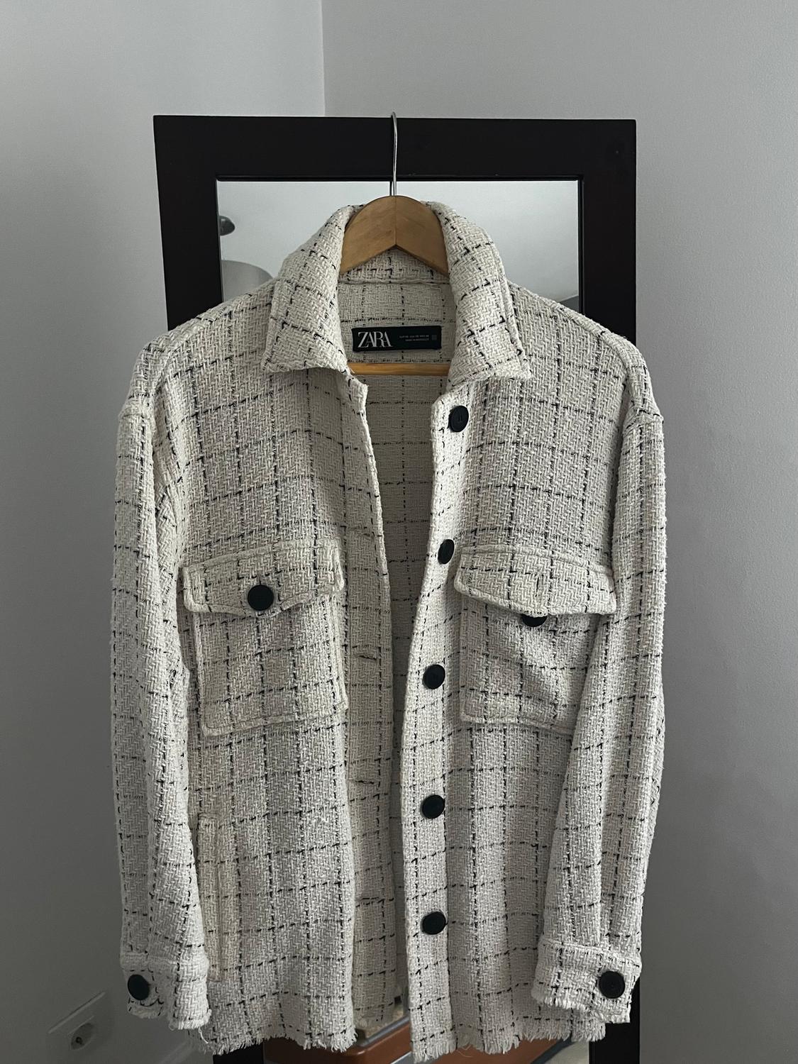 White textured fabric jacket with black buttons and pockets, hanging on a hanger in front of a mirror. Visible label: Zara.