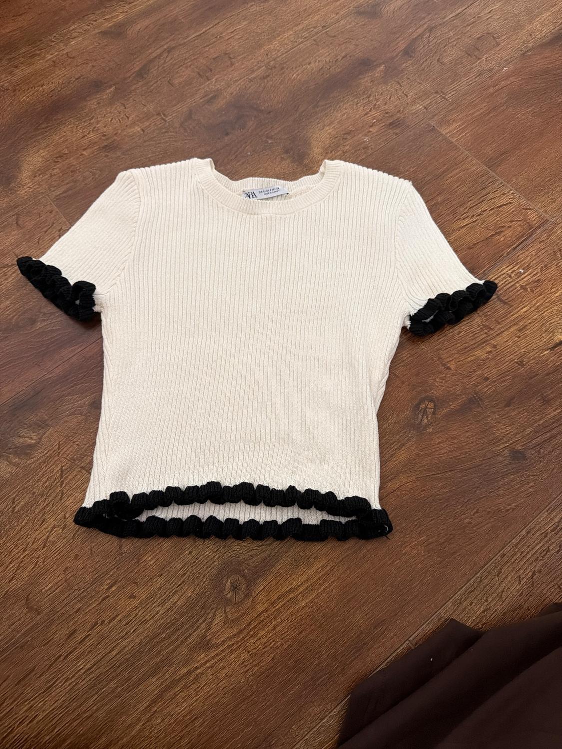 White knitted T-shirt with black frills on sleeves and hem, on a brown wooden floor. Visible label on the collar.