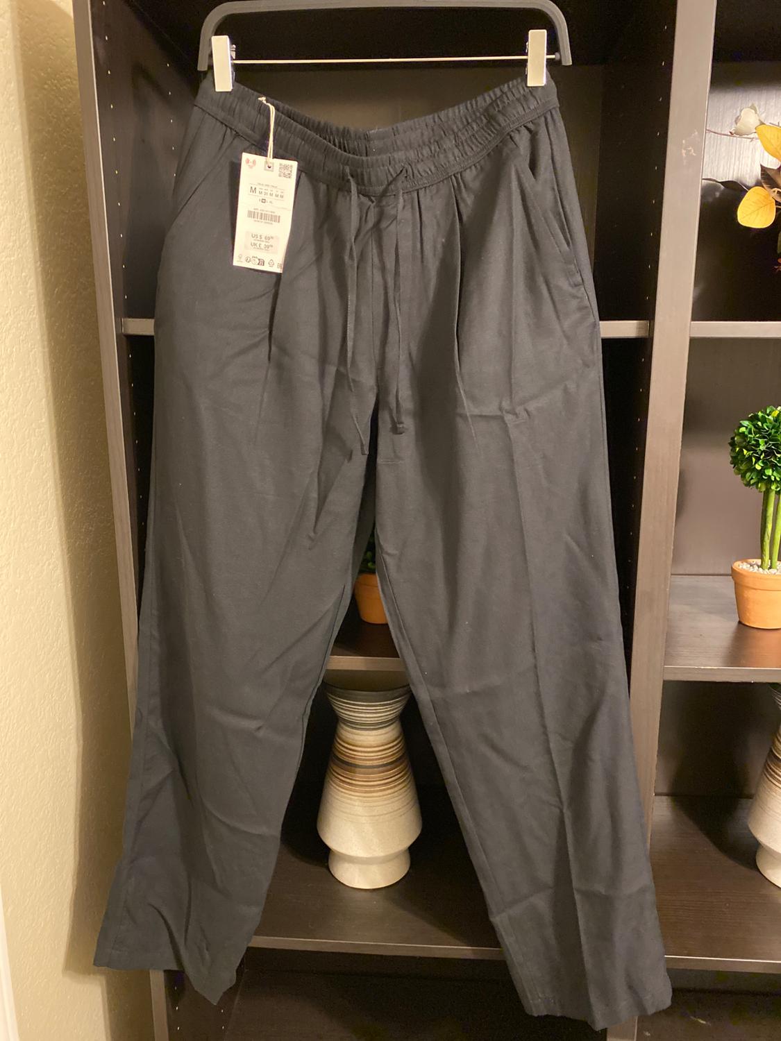 Black trousers hanging on a hanger with a visible tag, against a dark shelf background with decorative plants.