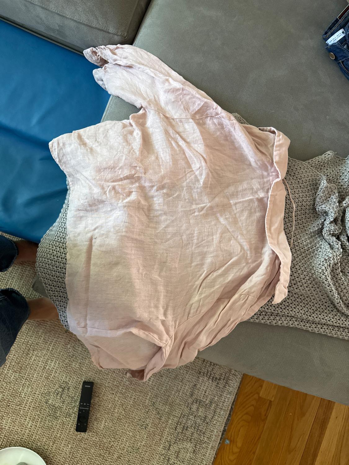 Light pink wrinkled shirt on a gray sofa, next to a knitted blanket and a black remote control on the wooden floor.