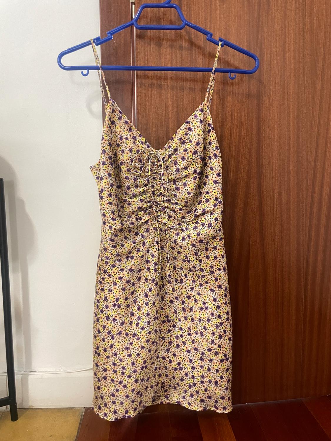 Yellow and purple floral print short strap dress. Front ruching with tie. Hanging on a blue hanger on a wooden door.