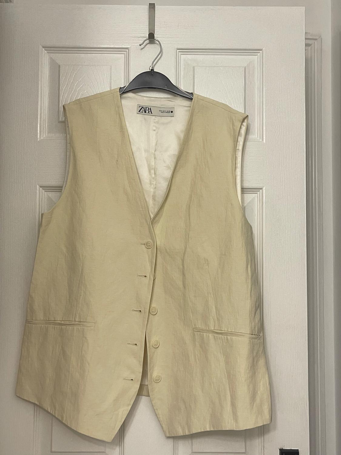 Beige waistcoat hanging on a black hanger over a white door. Visible label with the text "ZARA".