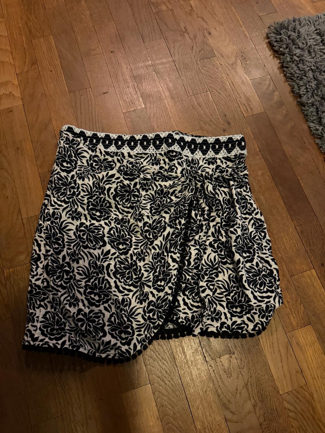 Short wrap skirt with a black-and-white floral print, decorated waistband and pom-pom trim, on a wooden floor.