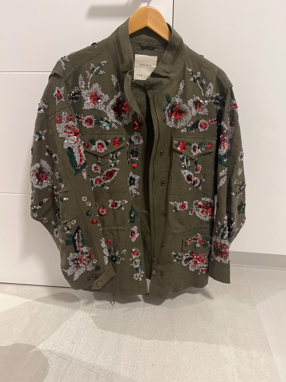 Olive green jacket with floral embroidery in red, white, and green, hanging on a wooden hanger against a white background.