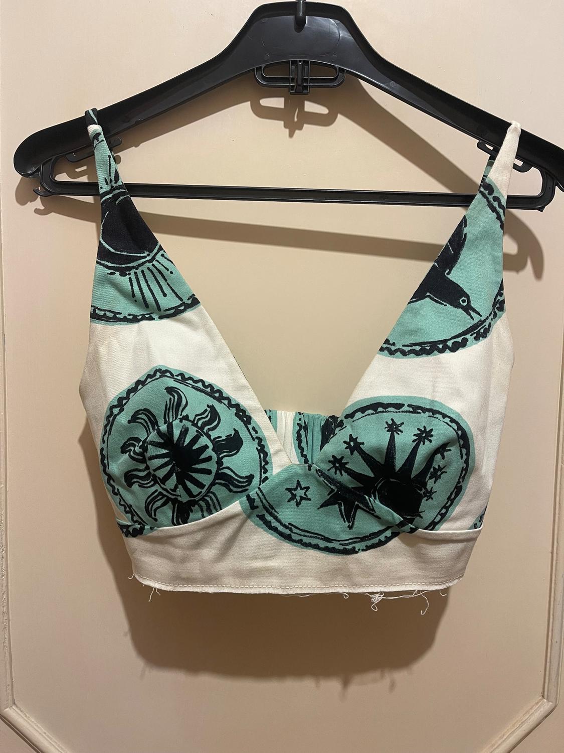 Cropped top with straps, sun and star print in green and black, hanging on a black hanger against a beige background.