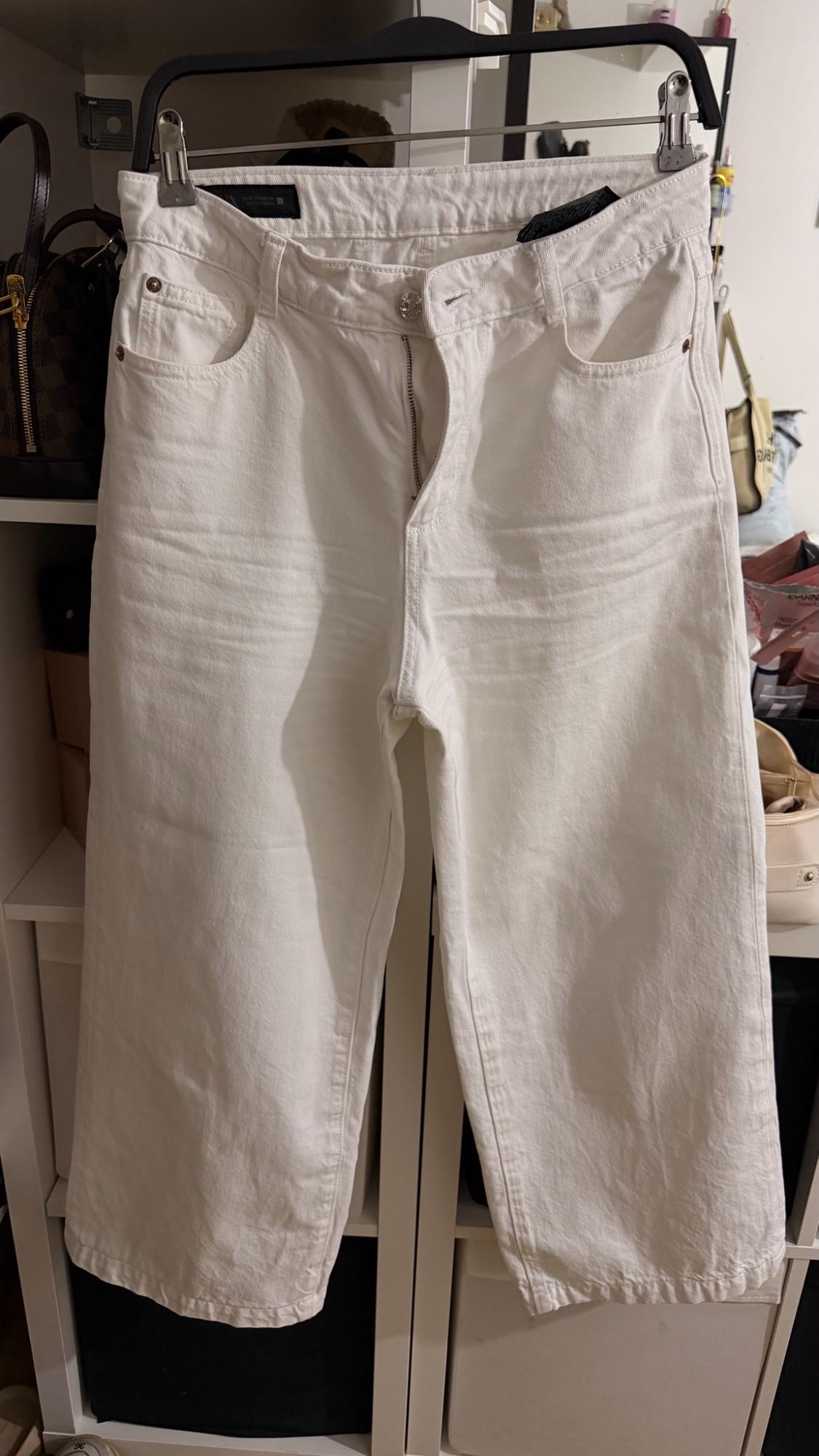 White jeans hanging on a hanger in a walk-in wardrobe. Relaxed atmosphere with white shelving, handbags and boxes in the background.