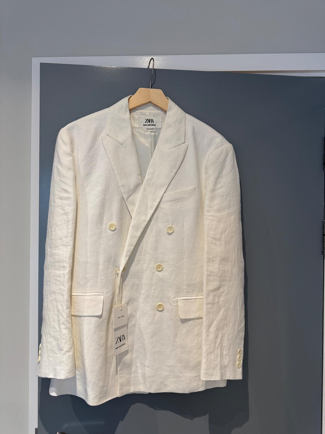 White cross-branded americana hung on a grey door. Visible labels with the text ZARA MAN SARTORIAL and 100% LINEN.