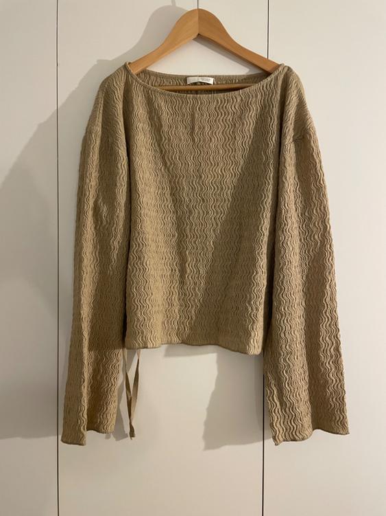 Beige sweater with a wavy texture, long sleeves, and a round neck, hanging on a wooden hanger against a white background.