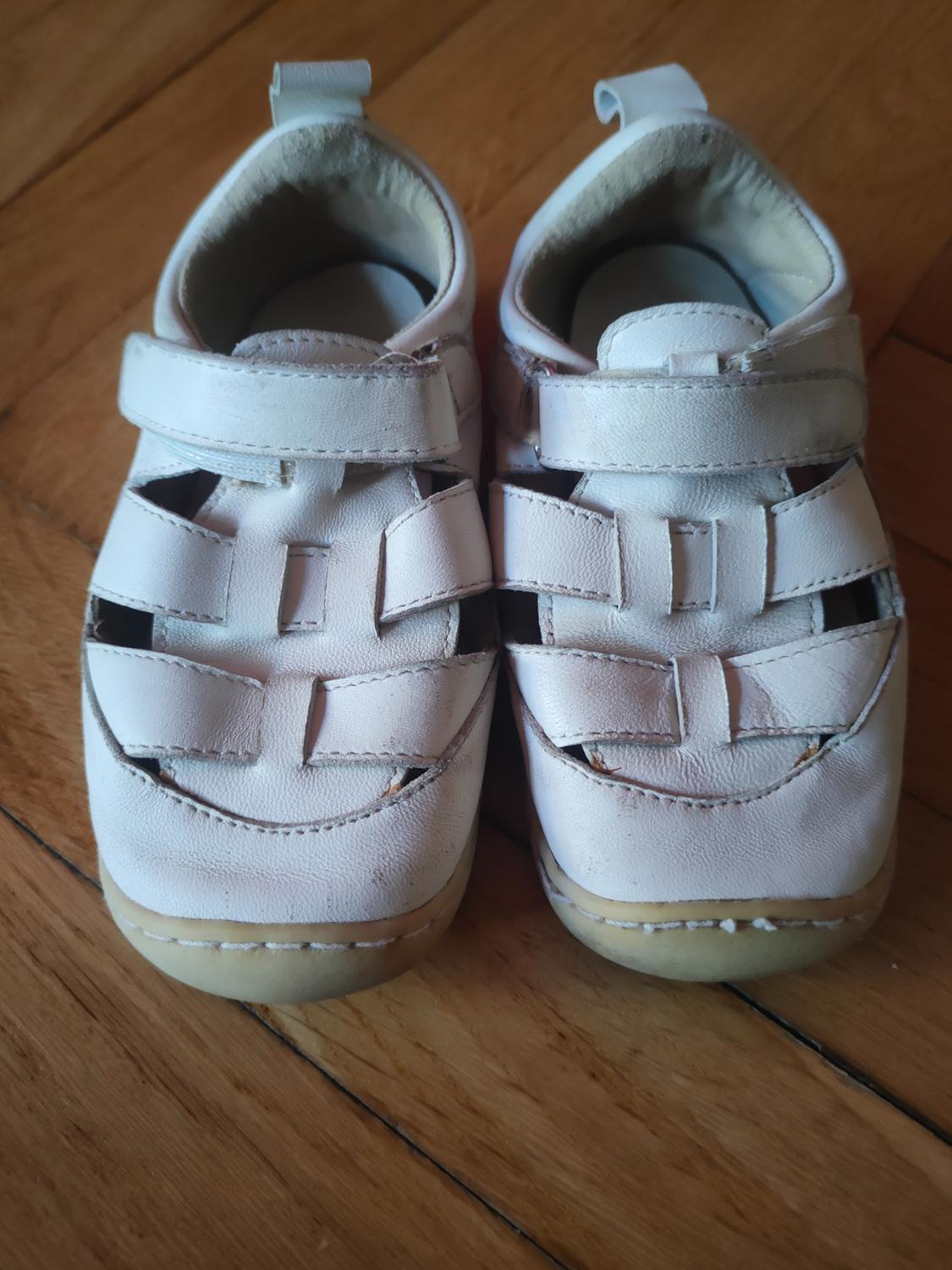 Pair of closed white sandals with an adjustable fastening and interwoven strap details, on a wooden floor.