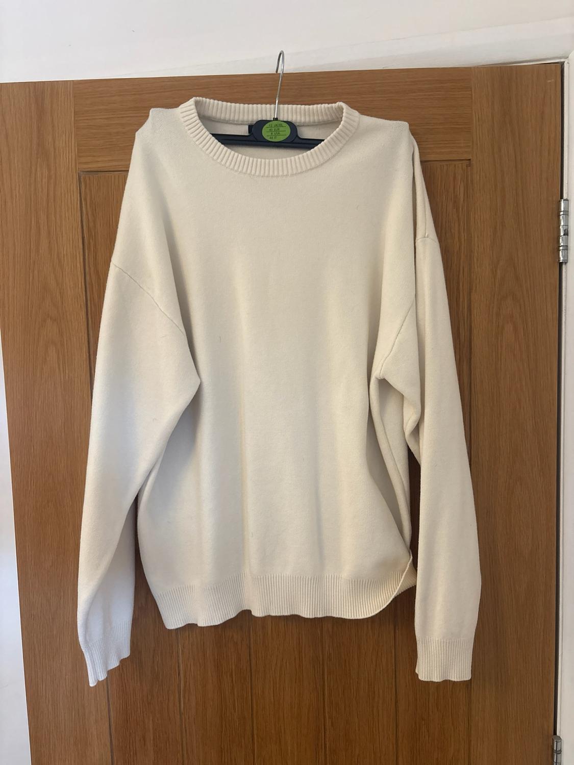 Cream crew-neck long-sleeved knitted jumper, hanging on a hanger over a wooden door.