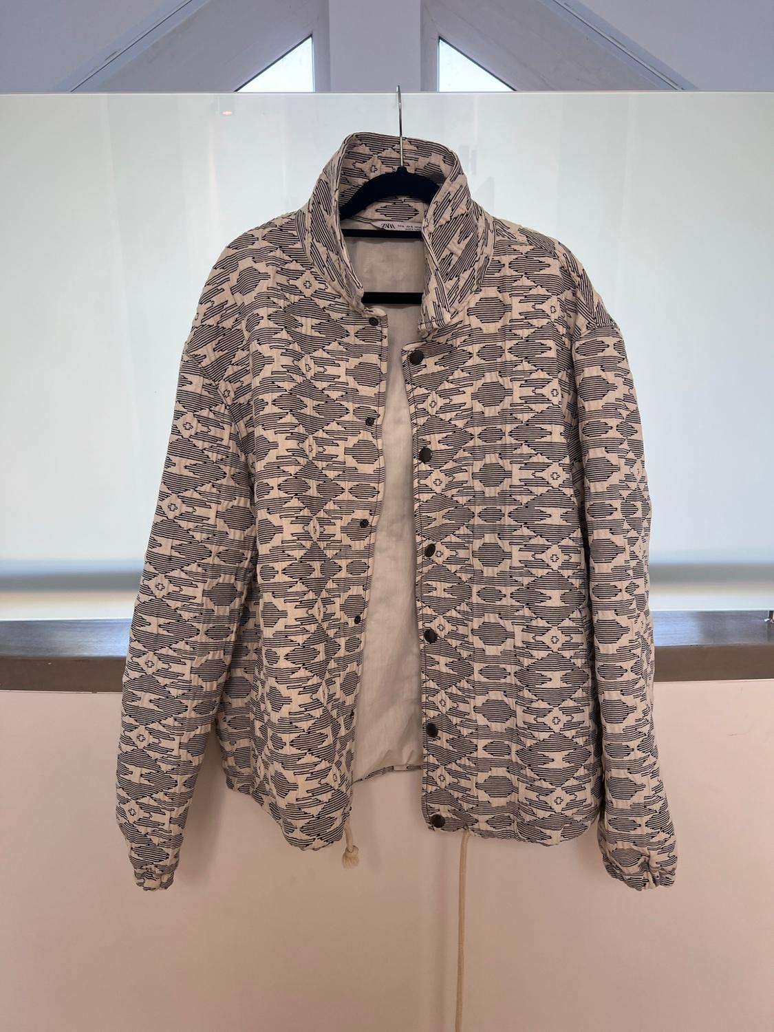 Padded jacket with a geometric print in neutral tones, hanging on a hanger in front of a window.
