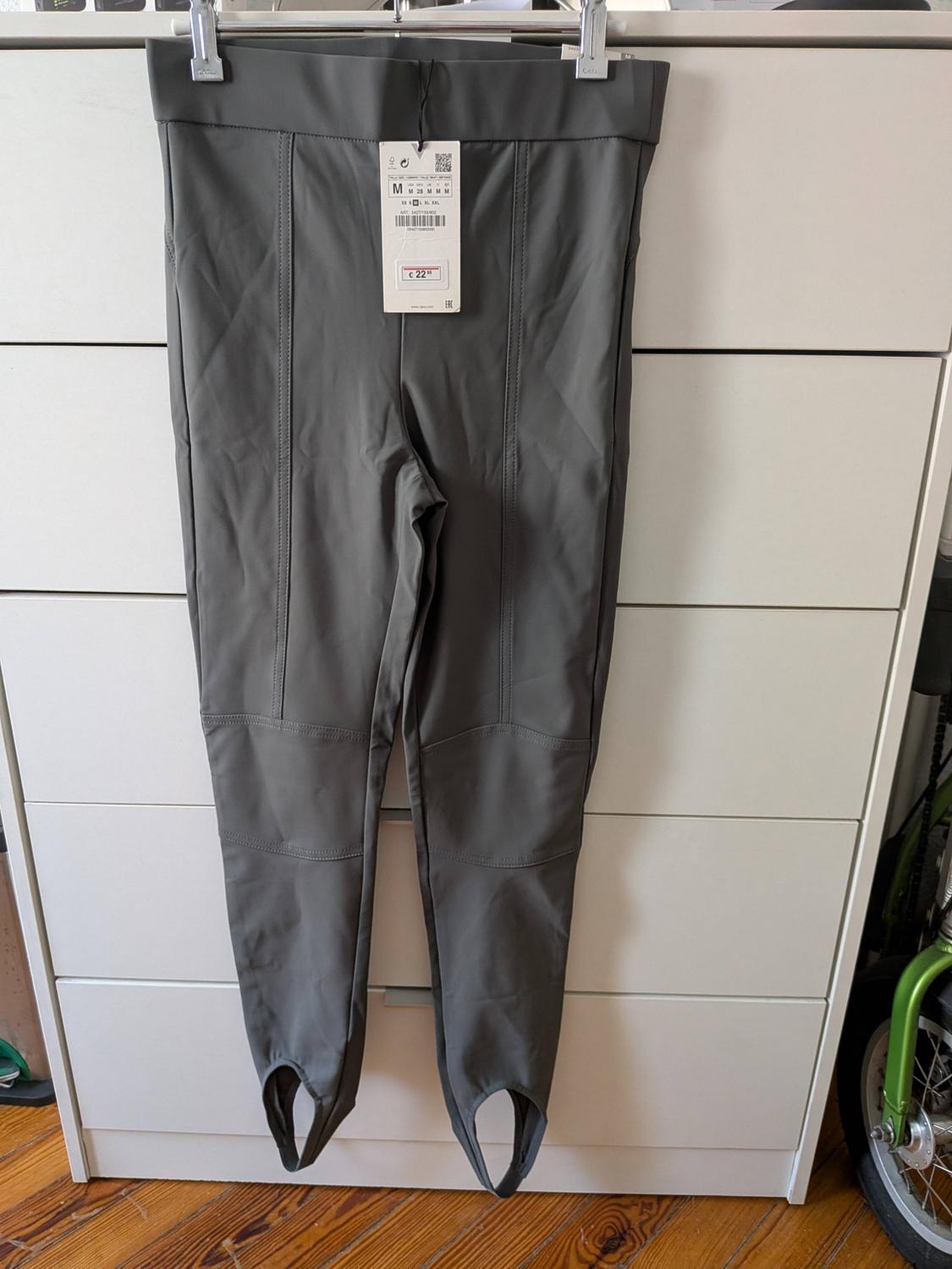 Grey stirrup trousers with front seams hanging on a hanger in front of a white chest of drawers. Label with text M and €22.95.