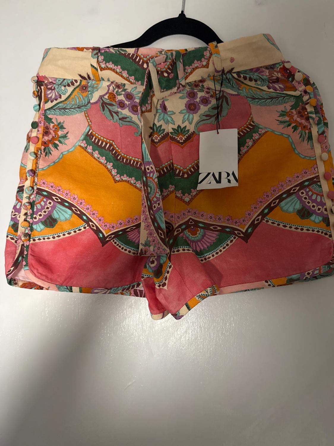 Colorful shorts with floral and geometric print in pink, orange, and green tones, hanging on a black hanger. Visible tag.