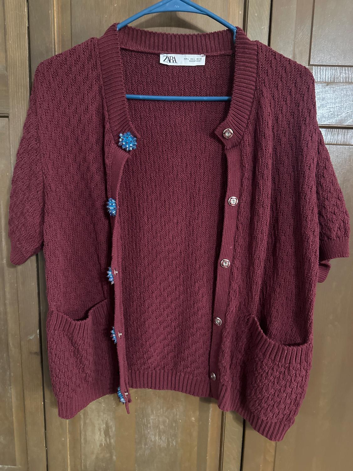 Burgundy short-sleeved cardigan with buttons and blue details, hanging on a blue hanger in front of a wooden door.
