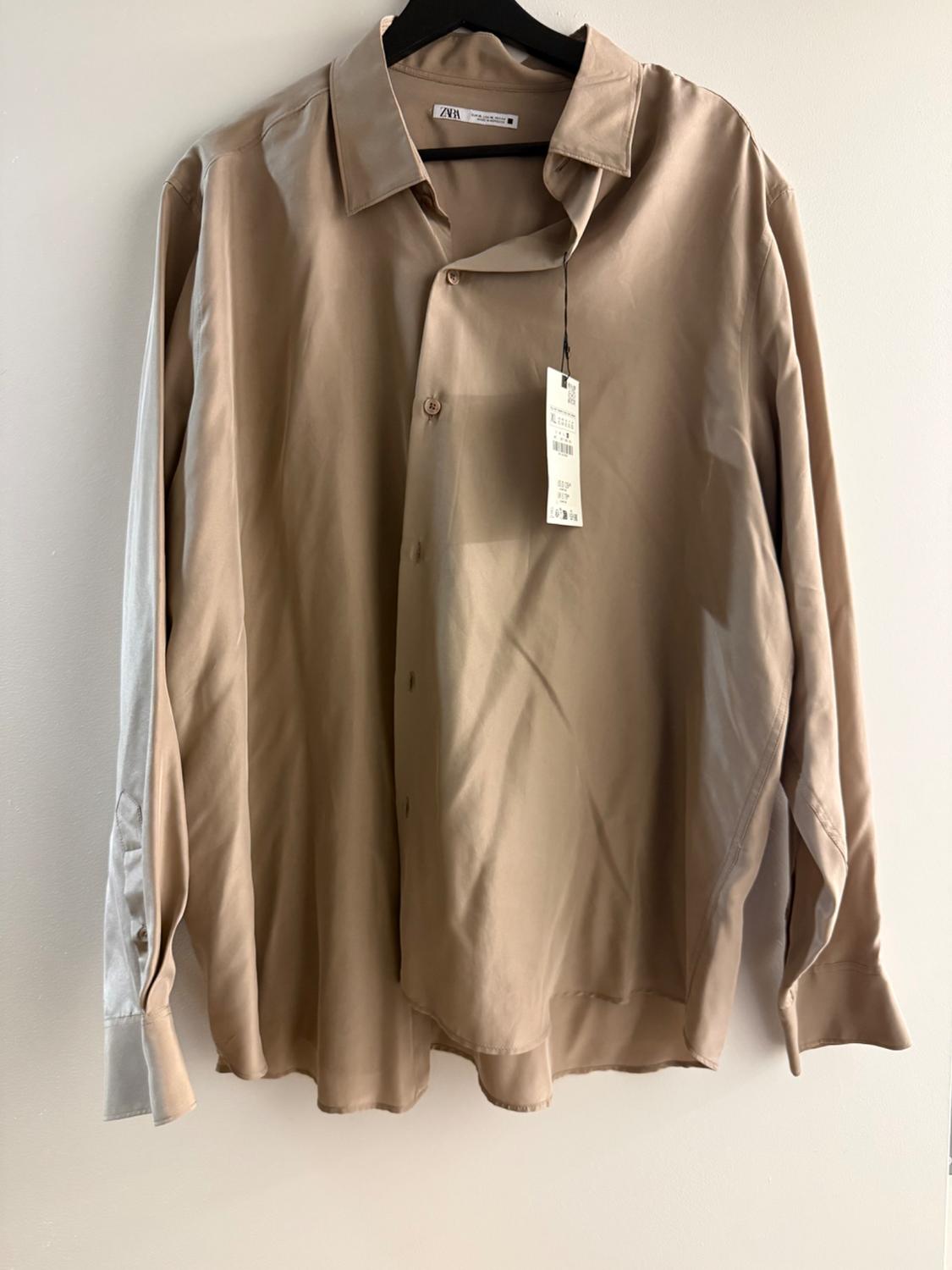Beige long-sleeved shirt hanging on a black hanger, with a visible label. Plain white background.