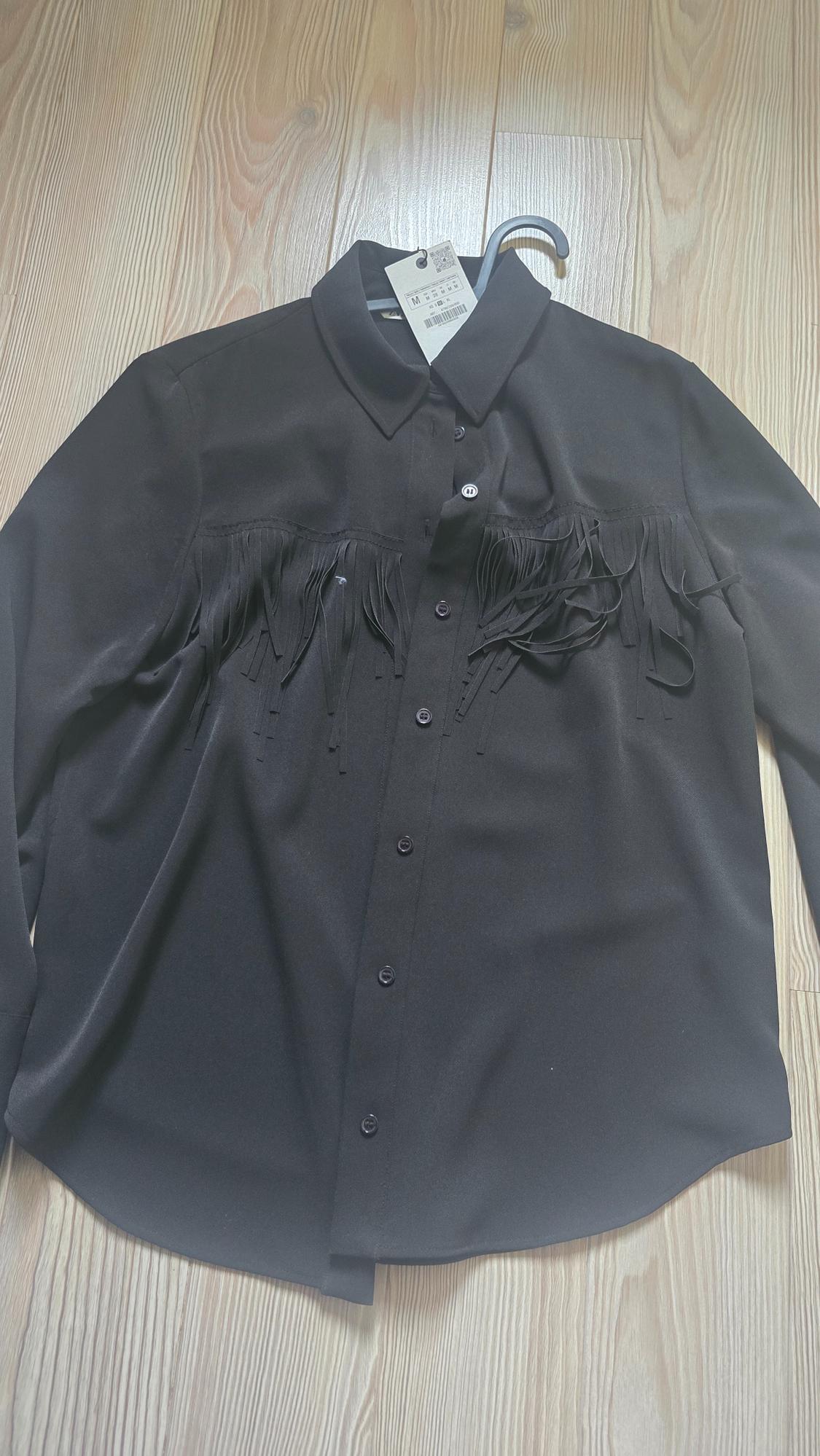 Black shirt with fringing on the chest, hanging on a hanger over a light wooden floor. Visible label with size M.