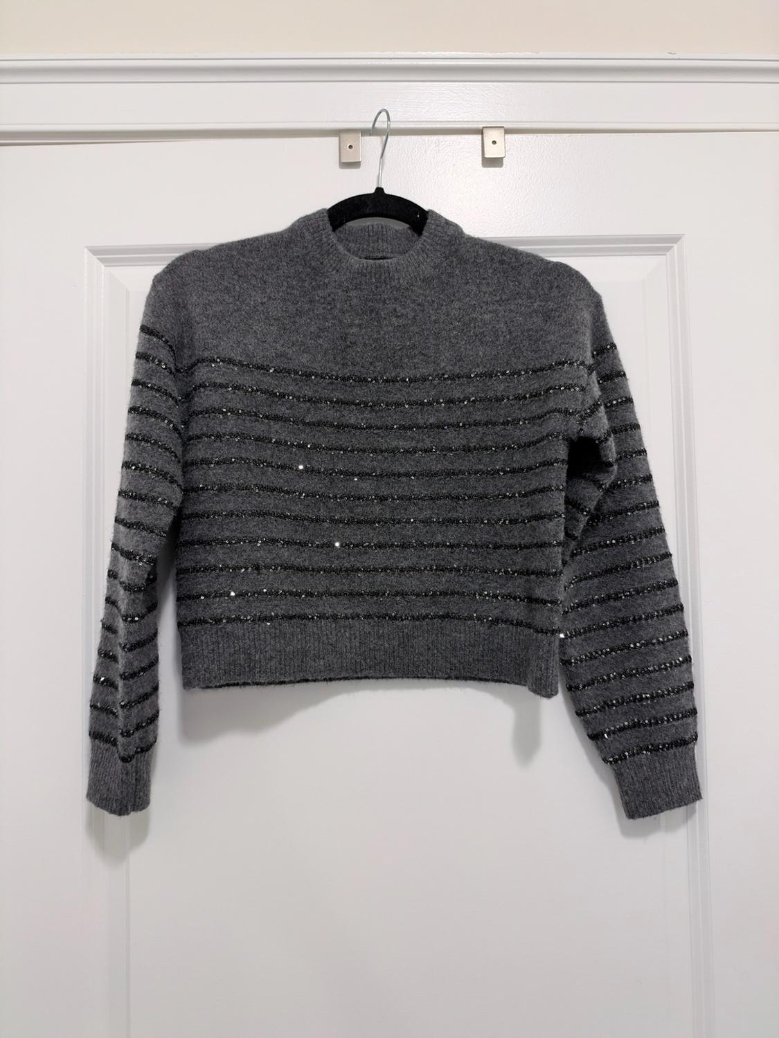 Gray knit sweater with a crew neck and shiny black horizontal stripes, hanging on a hanger over a white door.