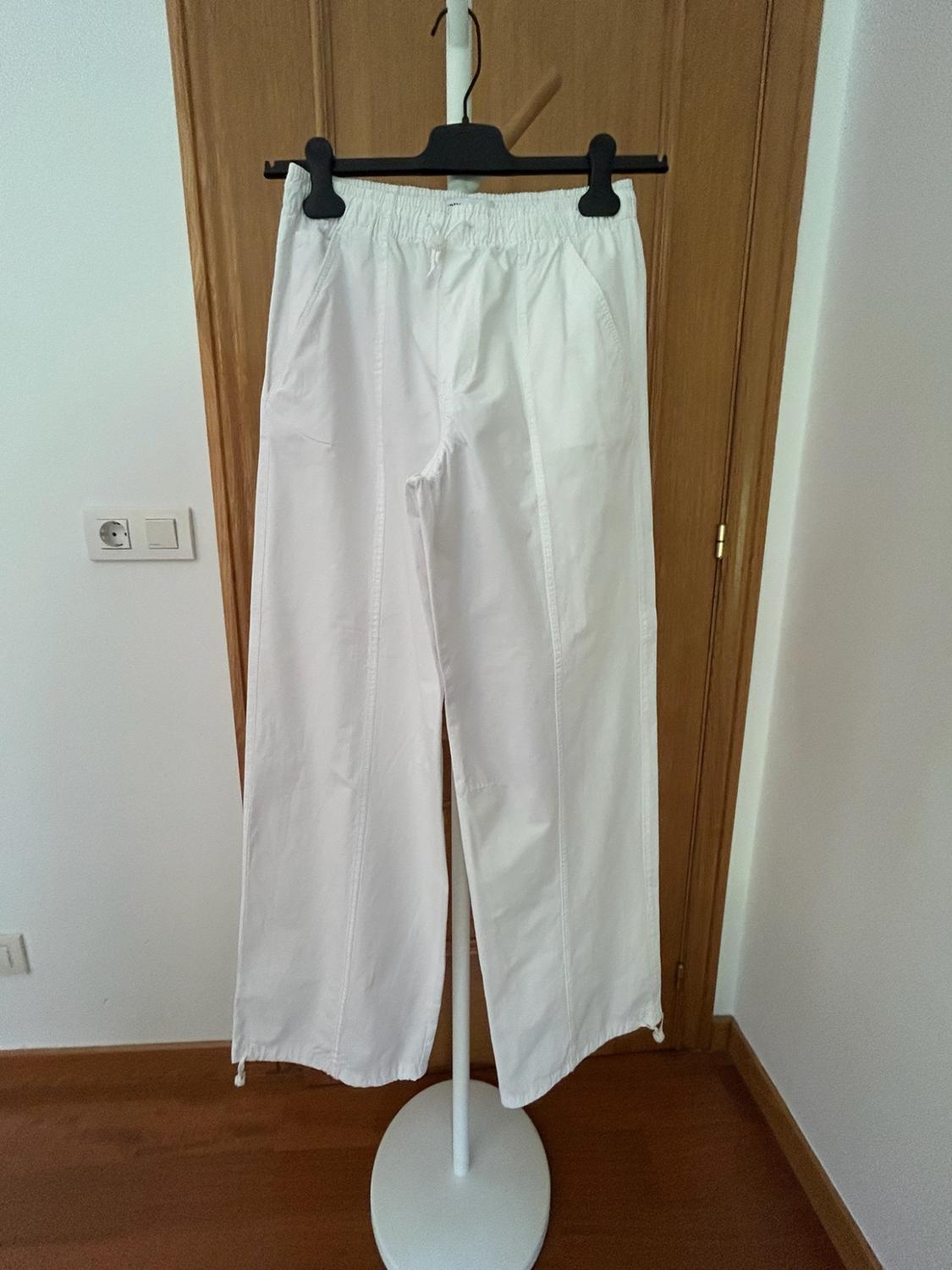White wide-leg trousers with an elasticated waist, hanging on a stand in front of a wooden door in a bright interior.
